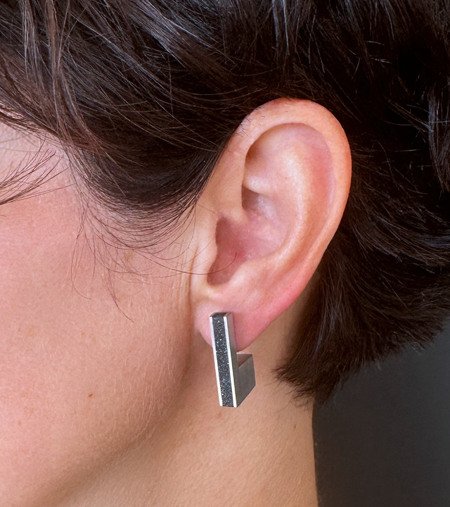 A close-up image showing a modern geometric stainless steel stud earring with a concrete-and-diamond dust inlay, worn on the ear and inspired by the minimalist Bauhaus design principles.