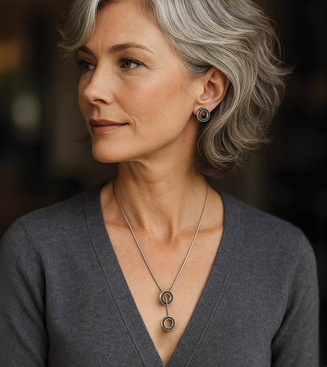 Elegant woman with silver-gray hair wearing contemporary minimalist jewelry. Her circular stainless steel and concrete earrings with diamond dust infusion complement her statement necklace. The pendant features two concrete-and-steel circular elements connected by a sleek stainless steel snakechain.