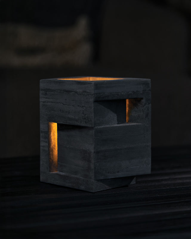 an architectural tealight holder A bold vertical form rises from its stepped base, pierced by precise openings that emit light from within. This contemporary concrete vessel stands sentinel-like, its strong geometric presence softened by the warm glow through its strategic apertures.
