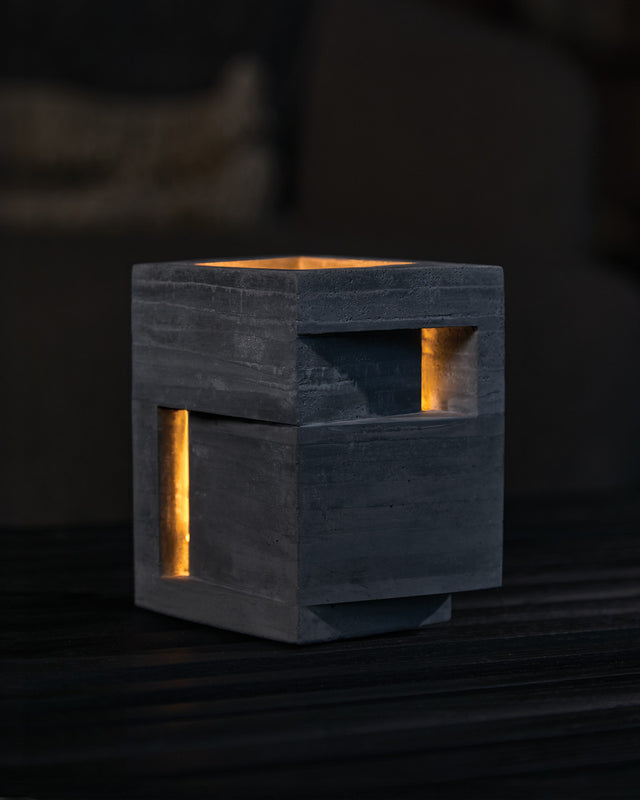 A charcoal-colored concrete candle holder with a geometric design, featuring rectangular cut-outs on each side. The holder is illuminated from within, creating a warm glow.