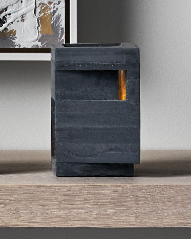 A bold vertical form rises from its stepped base, pierced by precise openings that emit light from within. This contemporary concrete vessel stands sentinel-like, its strong geometric presence softened by the warm glow through its strategic apertures. architectural tealight holder