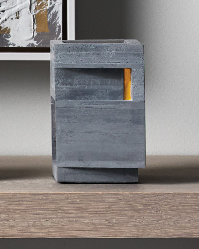 A bold vertical form rises from its stepped base, pierced by precise openings that emit light from within. This contemporary concrete vessel stands sentinel-like, its strong geometric presence softened by the warm glow through its strategic apertures.