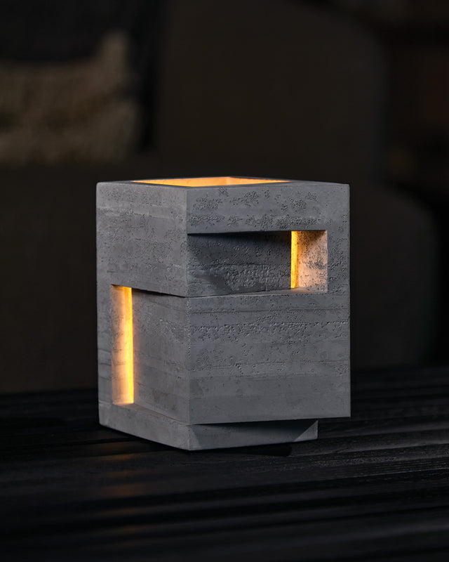 A bold vertical form rises from its stepped base, pierced by precise openings that emit light from within. This contemporary architectural concrete vessel stands sentinel-like, its strong geometric presence softened by the warm glow through its strategic apertures. Household by KONZUK