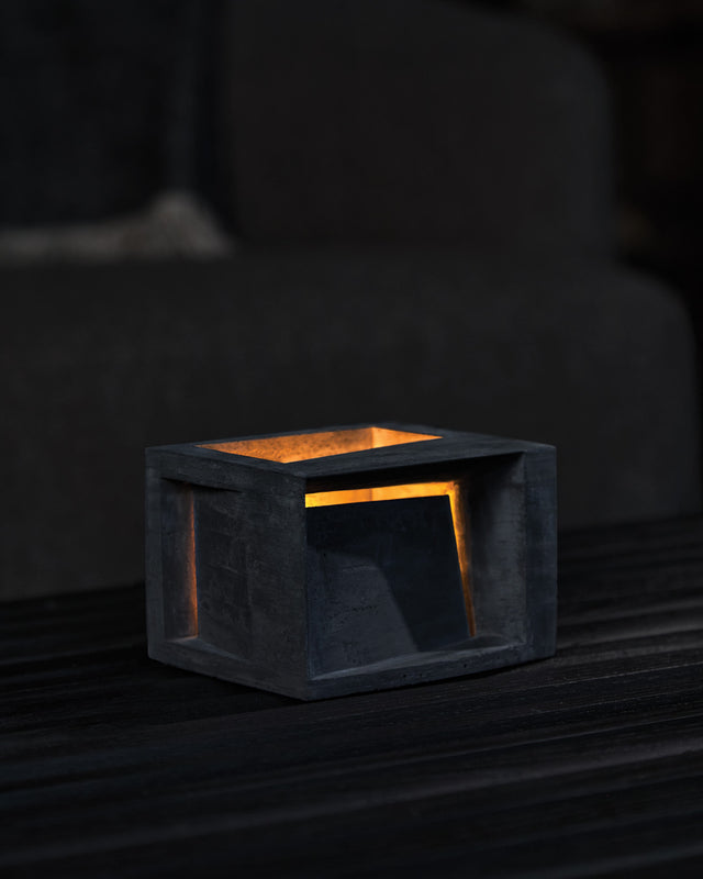 A black, landscape-oriented concrete form with a textured surface, displaying an illuminated tealight candle inside.