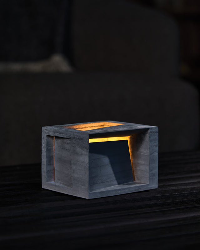 A landscape-oriented concrete form with a charcoal color, featuring an illuminated interior visible through a cut-out front facade, placed on a wooden surface.