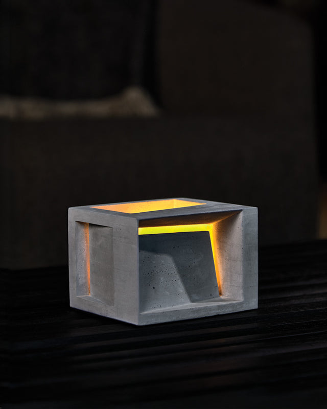 A concrete tealight candle holder in a natural color, with an illuminated interior visible through an opening in the front facade, placed on a dark surface.