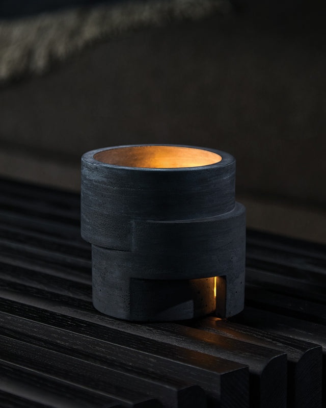 A black, textured concrete candle holder with a protective cork base, featuring a curved aperture and a small passageway, displaying a lit candle inside.