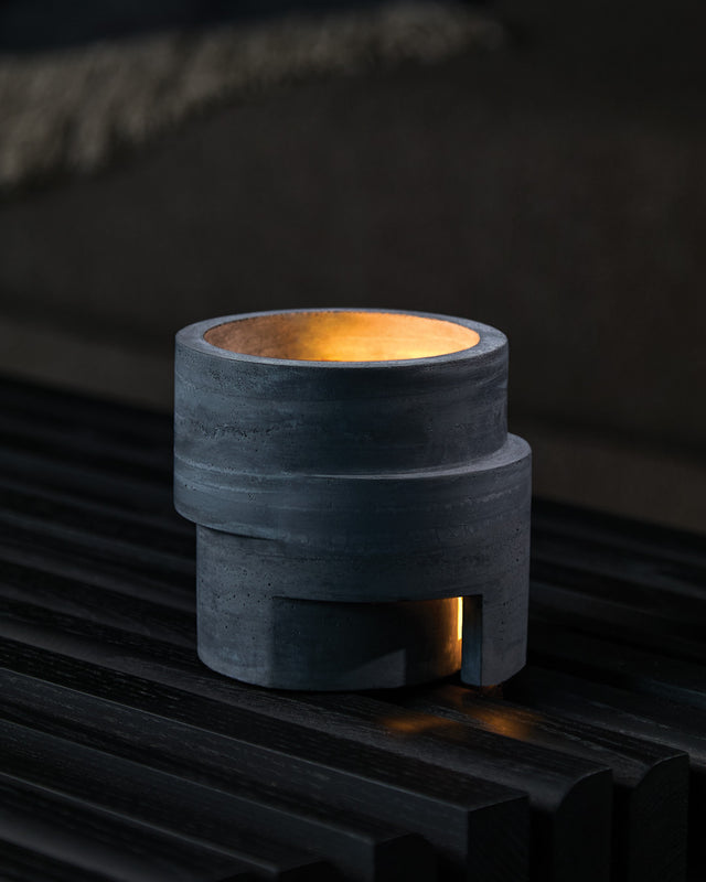 A charcoal-colored concrete candle holder with a stepped circular design, housing a lit tealight candle that casts light through the aperture. rotunda Habitat KONZUK