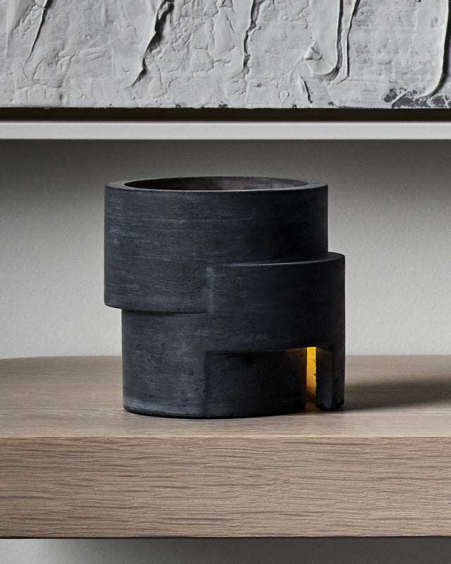 The Rotunda is a concrete tea light holder featuring bold cylindrical forms cast in tiered layers, inspired by classical architectural design. With its distinctive circular opening at the top and a rectangular window at the base, it creates a striking vertical play of light that radiates warm amber tones both upward and outward when lit. This sculptural piece brings monumental presence and sophisticated ambiance to your space, transforming any room into a gallery-like environment with its dramatic shadows a