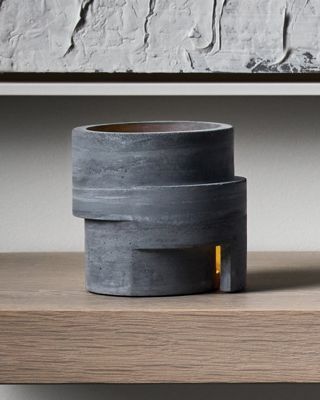 The Rotunda is a concrete tea light holder featuring bold cylindrical forms cast in tiered layers, inspired by classical architectural design. With its distinctive circular opening at the top and a rectangular window at the base, it creates a striking vertical play of light that radiates warm amber tones both upward and outward when lit. This sculptural piece brings monumental presence and sophisticated ambiance to your space, transforming any room into a gallery-like environment with its dramatic shadows a