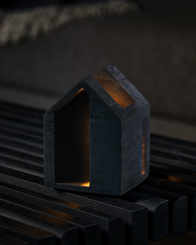 A black textured concrete candle holder with a cork base, displaying a lit tealight candle inside, placed on a wooden surface.