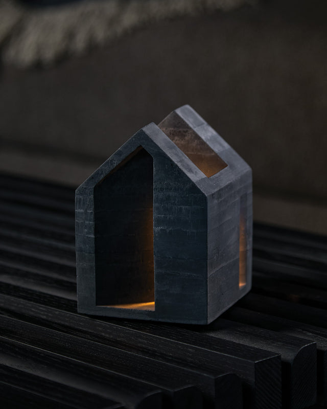 A hand-cast concrete candle holder in a house shape with a cork base, illuminated from the inside, on a wooden surface.