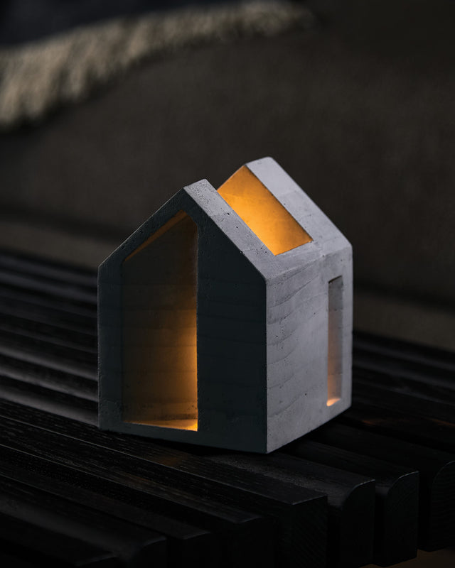 A textured concrete candle holder in a house shape with a cork base, illuminated from the inside with a warm light