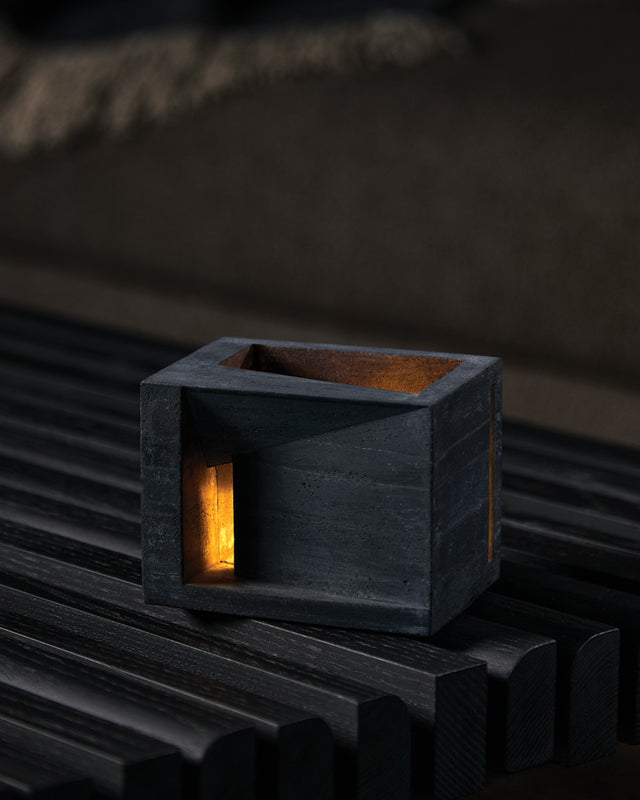 A black concrete candle holder with a textured finish, placed on a wooden surface, with a single-side aperture and illuminated interior visible. Villa tealight holder from KONZUK Habitat series