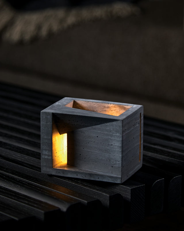 A hand-cast concrete decorative object with a charcoal color, featuring a minimalist design with converging angular planes, and a lit candle inside providing a warm glow.