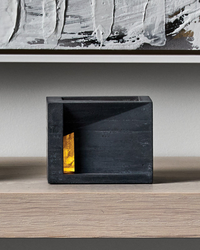 A black concrete candle holder with a textured finish, with a single-side aperture and illuminated interior visible. Villa tealight holder from KONZUK Habitat series