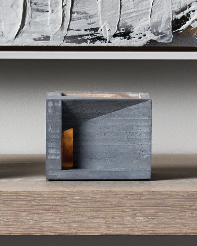 Villa is a concrete tea light holder that reveals the beautiful layered striations of cast concrete, like geological strata that tell the story of its creation. With its cubic architecture and generous rectangular opening, the candlelight illuminates these textured layers from within, offering an intimate glimpse into the imagined life of its inhabitant—as if peering through a window into a warm, lived-in space. This sculptural piece brings contemplative depth and serene ambiance to your home, transforming