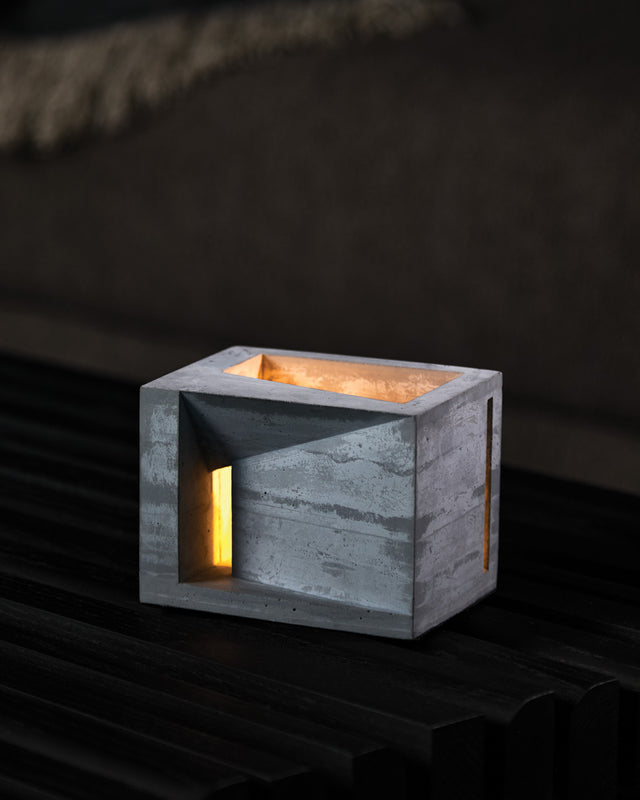 A minimalist architectural concrete form with a protective cork base, featuring a passage for light, illuminated from within. The form is displayed against a dark background on a wooden surface.