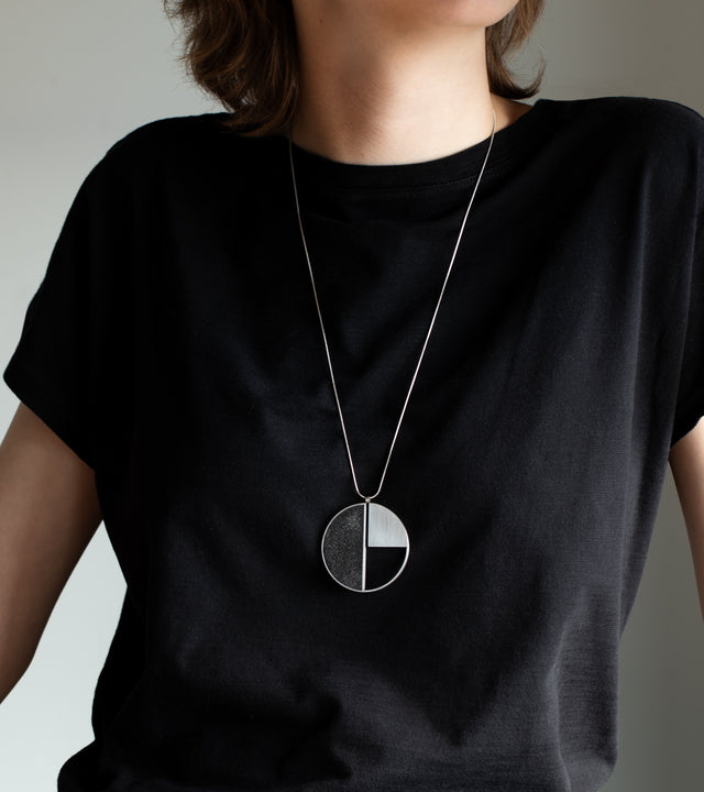 This striking geometric necklace pays homage to László Moholy-Nagy's pioneering Bauhaus aesthetic, featuring a bold circular pendant divided into contrasting quadrants of textured black and brushed silver. The modernist design captures Moholy-Nagy's exploration of light, material, and geometric abstraction, creating a wearable piece of art history. Suspended on a delicate silver chain, this statement piece brings avant-garde sophistication to any outfit while celebrating the enduring influence of constructi