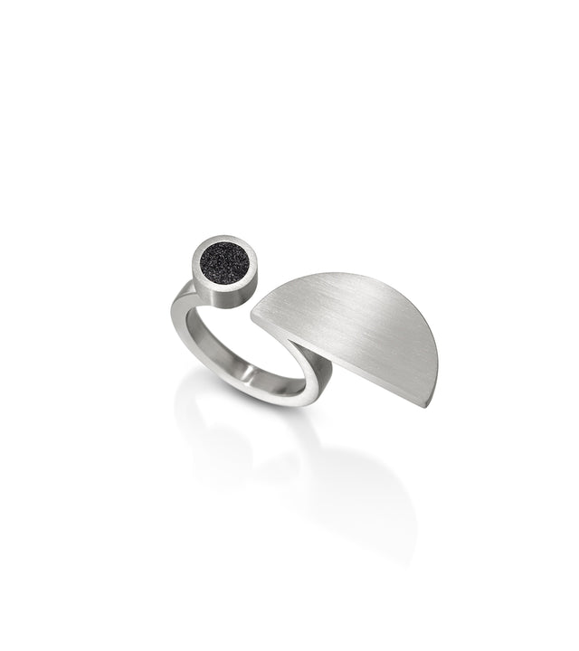 A contemporary ring with a geometric design, featuring a black diamond dust accent on a concrete surface, all in silver color.