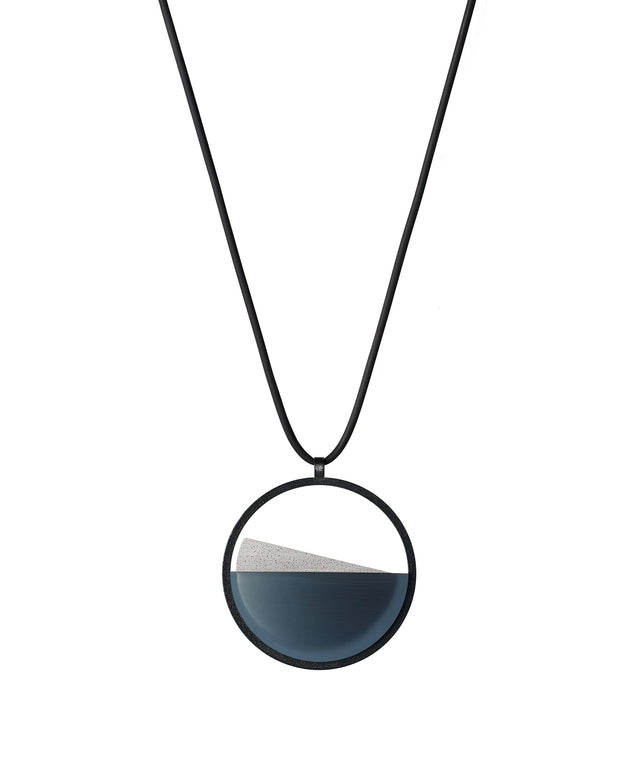 The Clara Small necklace in cerulean blue resin captures the essence of clarity in a refined, understated form. Named from the Latin clarus, meaning “bright” or “clear,” it reveals a quiet balance between resin and concrete. Within the circular frame, the concrete emerges gently from the resin’s surface, creating a soft meeting of tone and texture.
