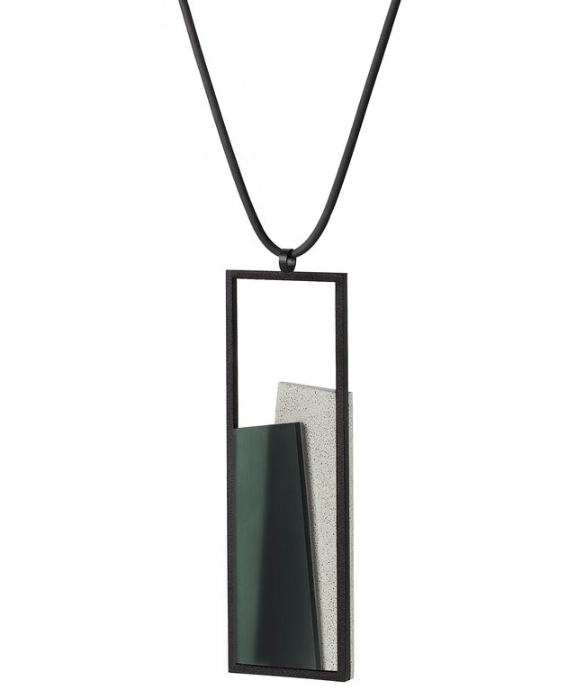 side view of The Eira necklace in Cypress Green brings together concrete and resin in a seamless composition that highlights material depth and subtle tone. Within the black rectangular frame, the concrete appears softly suspended in resin, giving the piece a quiet sense of balance and dimension. The surface catches light in a gentle, diffused way, creating an effect that feels both grounded and luminous. The Eira necklace offers a refined expression of modern minimalism.