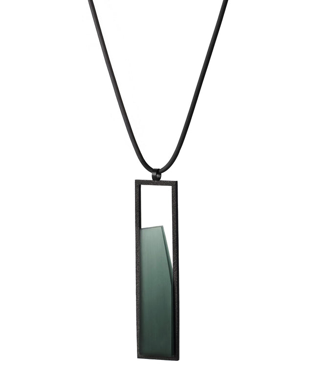 side view of The Lucia necklace in Cypress green resin features a sleek rectangular pendant that emphasizes form and material. A cast resin element rests within a black frame, creating a sense of depth and quiet luminosity. The elongated shape draws the eye, offering a refined balance between structure and softness. Minimal and striking, the Lucia necklace adds a sculptural accent that complements both everyday and statement looks.