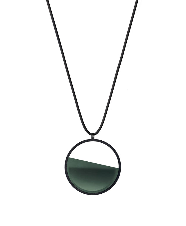 The Vestra Small necklace in Cypress green resin captures the same sense of balance and quiet strength in a more understated form that the larger version. Named from the Latin vestra, meaning “yours,” it reflects a personal connection between light, material, and wearer. The smaller circular frame holds a smooth resin plane with a cast concrete inset that reveals gentle depth and tone, creating a soft glow close to the body.
