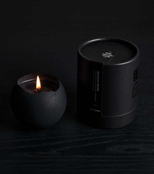 A black concrete candle with black wax, placed inside a black cylindrical vessel, accompanied by a small black box with a candle lid.