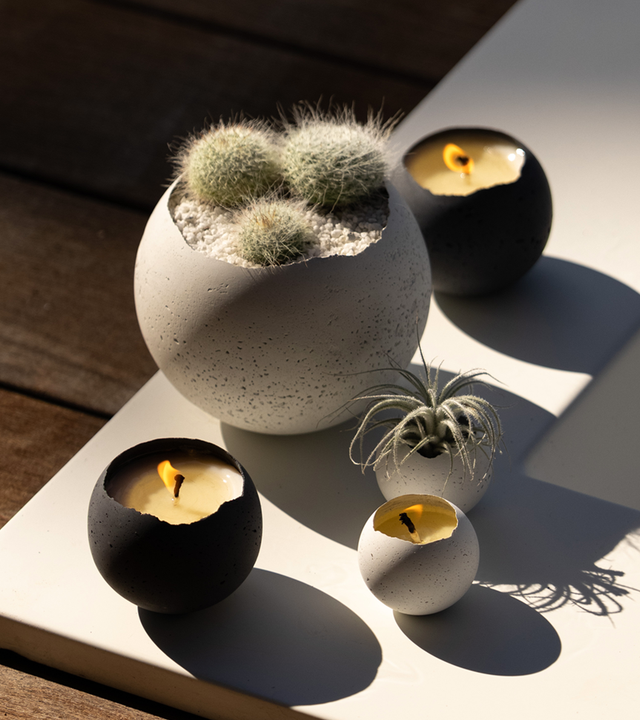A white concrete vessel with a pitted texture, displayed in an outdoor setting with a plant inside and candles around.