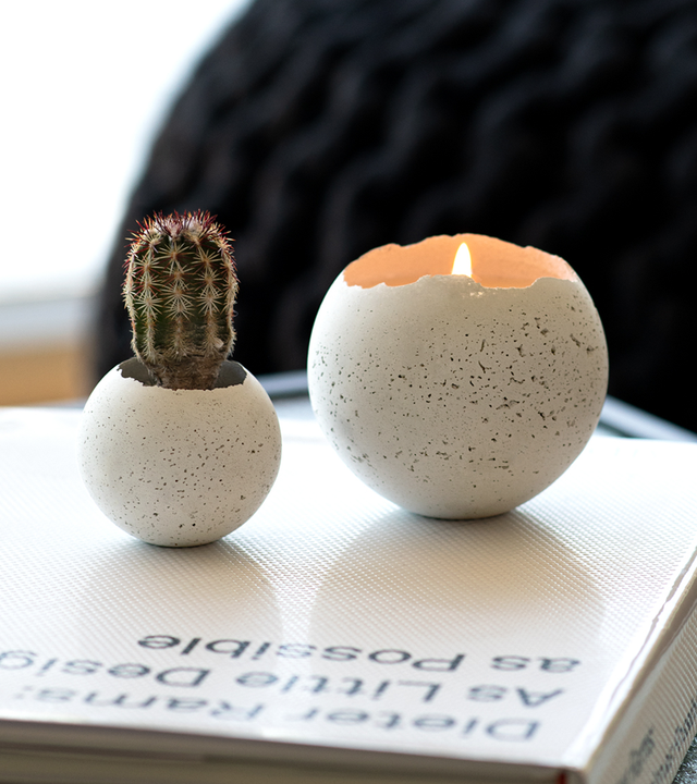 A hand-poured white-tinted concrete candle vessel with a burning candle inside, placed on an open book next to a cactus.