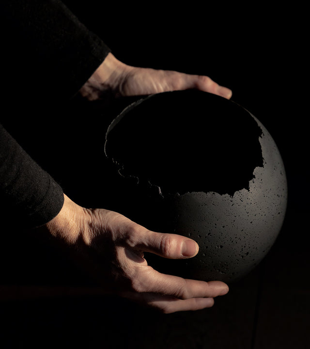 Hand-crafted black concrete vessel with a live-edge opening, designed for use as a planter. being held
