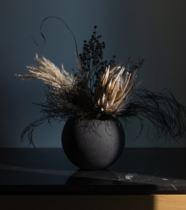 A charcoal-tinted concrete vessel with a live-edge opening, filled with dry plants and decorative foliage, placed on a dark surface against a dark background.