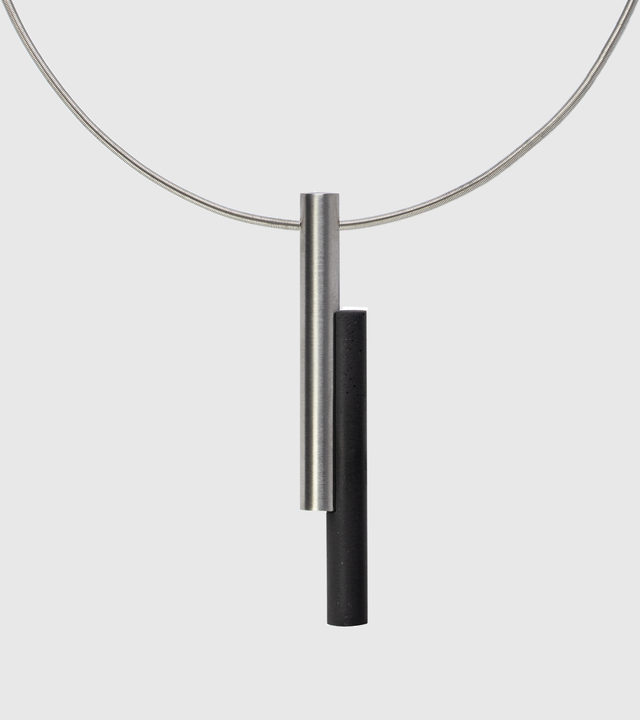 A necklace featuring a concrete cylinder and a brushed stainless steel cylinder, hanging from a stainless steel cable.