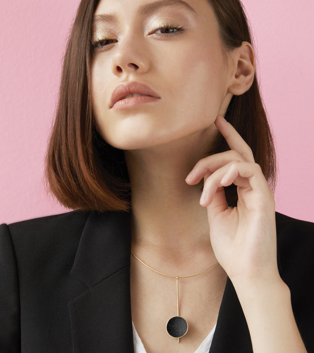 This striking Inspira necklace features a diamond dust-encrusted concrete disc set into luxurious 14 karat gold, named after the mysterious beauty of black holes. The elegant drop pendant is suspended on a sleek vertical bar and delicate omega chain, creating sophisticated movement while the sparkling texture catches light like distant stars. Timelessness is paired with modernist minimalism in this lightweight, statement-making piece that embodies cosmic wonder and transitions effortlessly from day to eveni