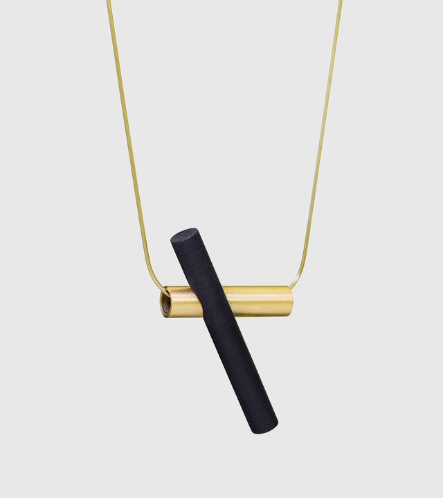 Suspended from a 14k gold snake-chain a black-tinted concrete cylinder cantilevers a brushed tube in solid 14k gold.