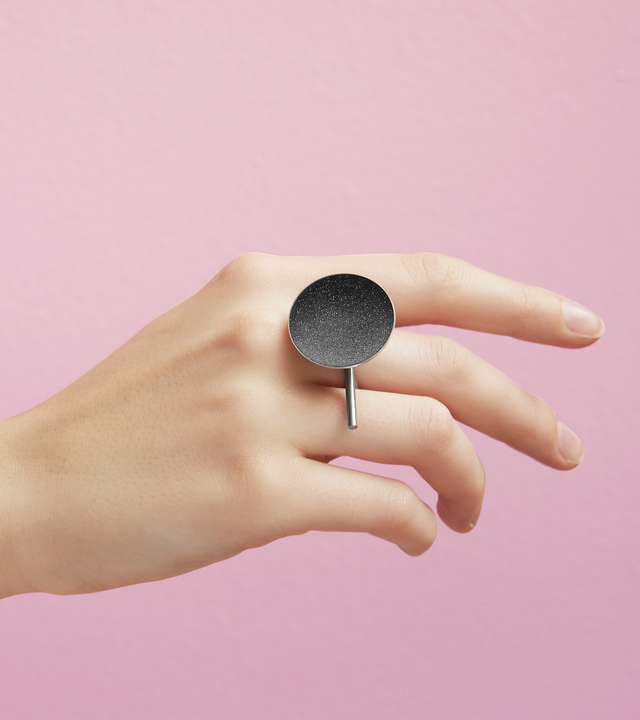 The Juno Major statement ring is features diamond dust infused into black concrete set into a small stainless steel dome architecturally connected to an extended horizontal post positioned onto a rounded steel frame.