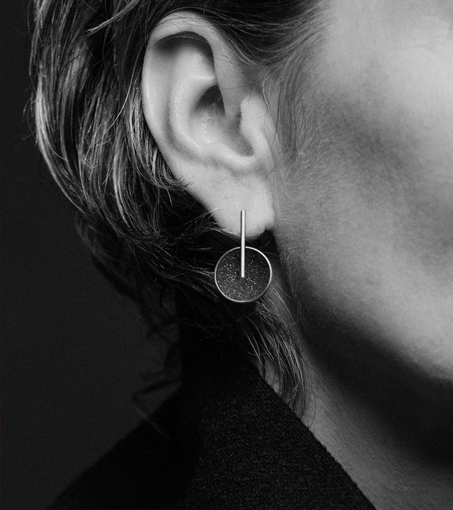 a model wears These striking minimalist earrings feature diamond dust-encrusted concrete domes set in stainless steel or 14k Ggold with a sleek geometric bar. The compact design offers comfortable all-day wear while the subtle sparkle of diamond dust against dark concrete adds refined texture that catches the light with every movement. Perfect for pairing with the matching pendant or wearing alone, these lightweight statement earrings bring contemporary artisan craftsmanship to any look from casual to elega