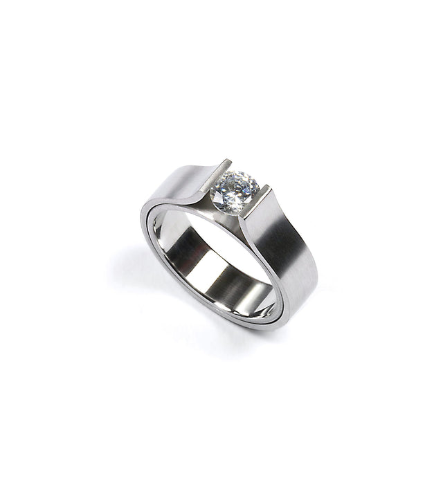 This ring showcases a striking minimalist design, featuring a half-carat diamond elegantly suspended within a sculptural band. Its clean, modern form highlights precision craftsmanship and architectural balance. Available in stainless steel, platinum, or a platinum and gold combination, this piece embodies refined simplicity and timeless strength.