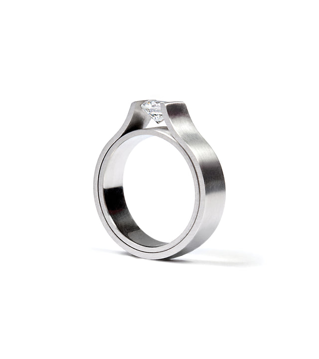 This ring showcases a striking minimalist design, featuring a half-carat diamond elegantly suspended within a sculptural band. Its clean, modern form highlights precision craftsmanship and architectural balance. Available in stainless steel, platinum, or a platinum and gold combination, this piece embodies refined simplicity and timeless strength.
