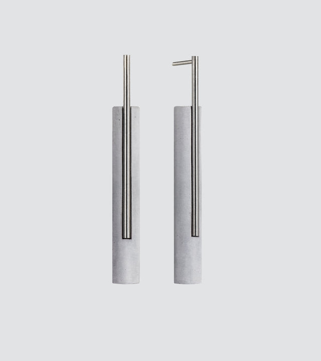 A pair of minimalist earrings featuring a brushed stainless steel rod in front of a small concrete cylinder, available in natural or black color.