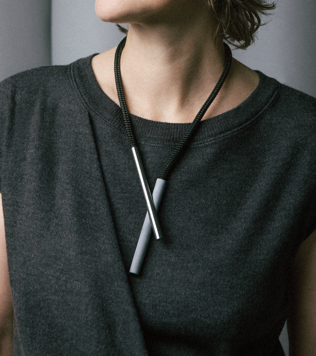 A person wearing a gray t-shirt is modeling a necklace with a concrete cylinder intersected by a stainless steel rod, connected by rare-earth magnets and hanging from a black nylon rope.
