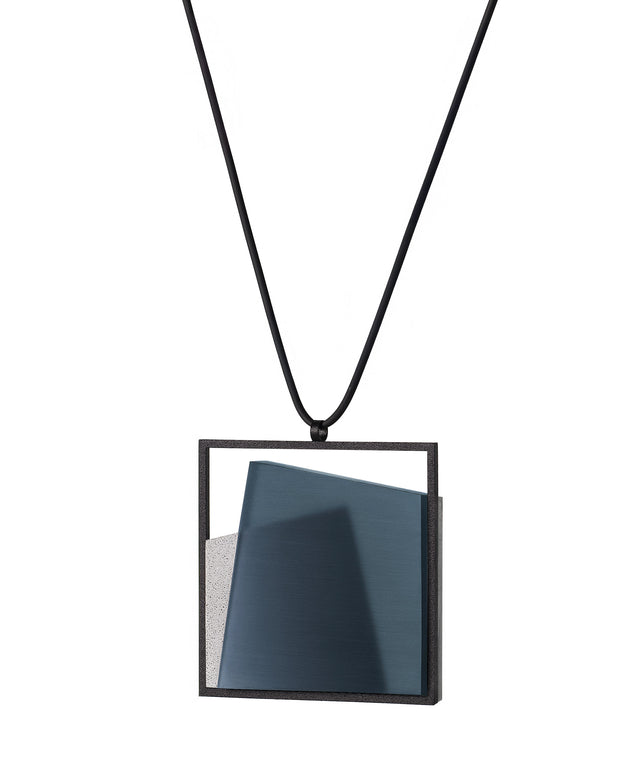 side view of The Astrid necklace in cerulean blue takes its name from Old Norse, meaning “divinely beautiful,” a reflection of its quiet, sculptural presence. Within the square frame, concrete intersects with resin in a deliberate composition that reveals depth and movement. The meeting of materials creates subtle shifts in tone and surface, drawing the eye to the balance between precision and softness. The Astrid necklace embodies refined minimalism with a sense of calm, architectural beauty.