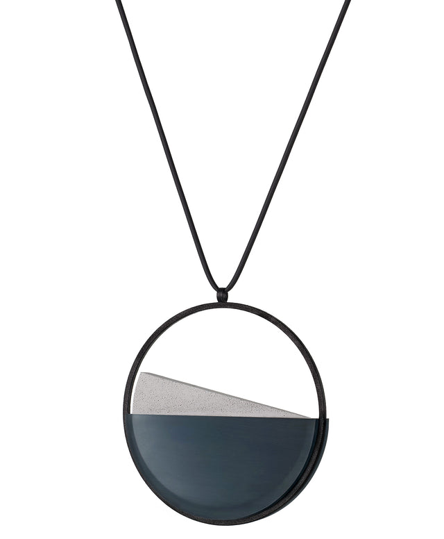 side view of The Clara necklace in cerulean blue embodies clarity and balance. Named from the Latin clarus, meaning “bright” or “clear,” it reflects the quiet harmony of light and material. Within the circular frame, concrete emerges from the surface of the resin, creating a gentle rise in tone and texture. The Clara necklace carries a calm, sculptural presence that feels both modern and enduring.