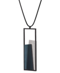 Side view of The Eira necklace in Cerulean Blue brings together concrete and resin in a seamless composition that highlights material depth and subtle tone. Within the black rectangular frame, the concrete appears softly suspended in resin, giving the piece a quiet sense of balance and dimension. The surface catches light in a gentle, diffused way, creating an effect that feels both grounded and luminous. The Eira necklace offers a refined expression of modern minimalism.