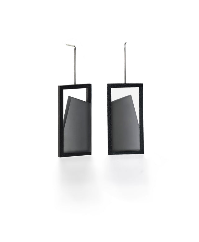 The Elena earrings play with light through a sleek, elongated form. A vertical resin plane reveals a smooth gradient that shifts from light to shadow, bringing subtle movement to the piece. The translucent resin captures and softens light, creating a quiet glow within the structured black frame. Modern and understated, these earrings offer a refined balance of geometry and fluid luminosity. in ash gray
