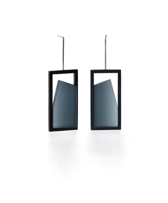 The Elena earrings play with light through a sleek, elongated form. A vertical resin plane reveals a smooth gradient that shifts from light to shadow, bringing subtle movement to the piece. The translucent resin captures and softens light, creating a quiet glow within the structured black frame. Modern and understated, these earrings offer a refined balance of geometry and fluid luminosity. in cerulean blue