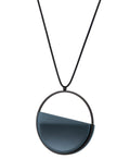 side view of The Vestra necklace in cerulean blue resin embodies balance and quiet strength. Its name, derived from the Latin vestra, meaning “yours,” speaks to a personal connection between wearer and form. Within the circular frame, concrete is cast into resin revealing a smooth plane of depth and tone, reflecting light in a soft, fluid way. The composition feels calm and grounded, offering a sculptural presence that becomes uniquely individual when worn.