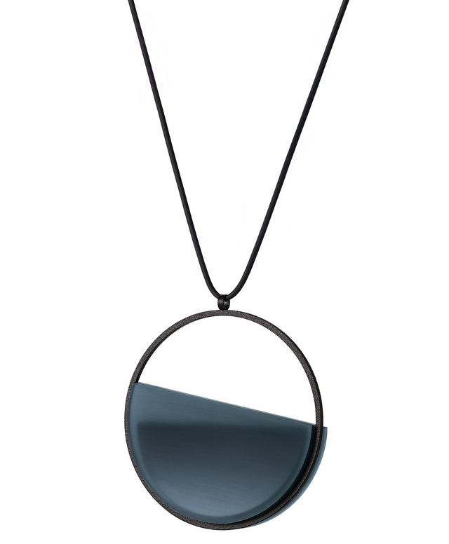 side view of The Vestra necklace in cerulean blue resin embodies balance and quiet strength. Its name, derived from the Latin vestra, meaning “yours,” speaks to a personal connection between wearer and form. Within the circular frame, concrete is cast into resin revealing a smooth plane of depth and tone, reflecting light in a soft, fluid way. The composition feels calm and grounded, offering a sculptural presence that becomes uniquely individual when worn.