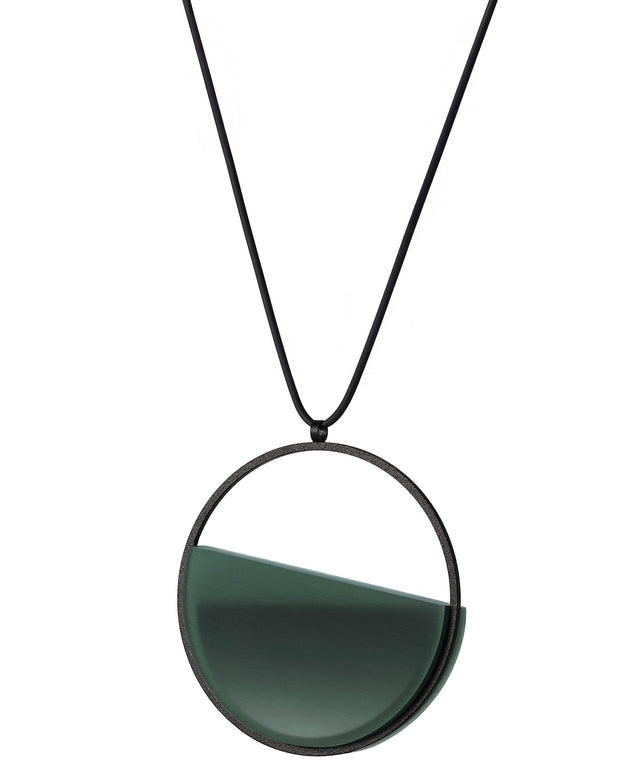 Side view of The Vestra necklace in Cypress green resin embodies balance and quiet strength. Its name, derived from the Latin vestra, meaning “yours,” speaks to a personal connection between wearer and form. Within the circular frame, concrete is cast into resin revealing a smooth plane of depth and tone, reflecting light in a soft, fluid way. The composition feels calm and grounded, offering a sculptural presence that becomes uniquely individual when worn.